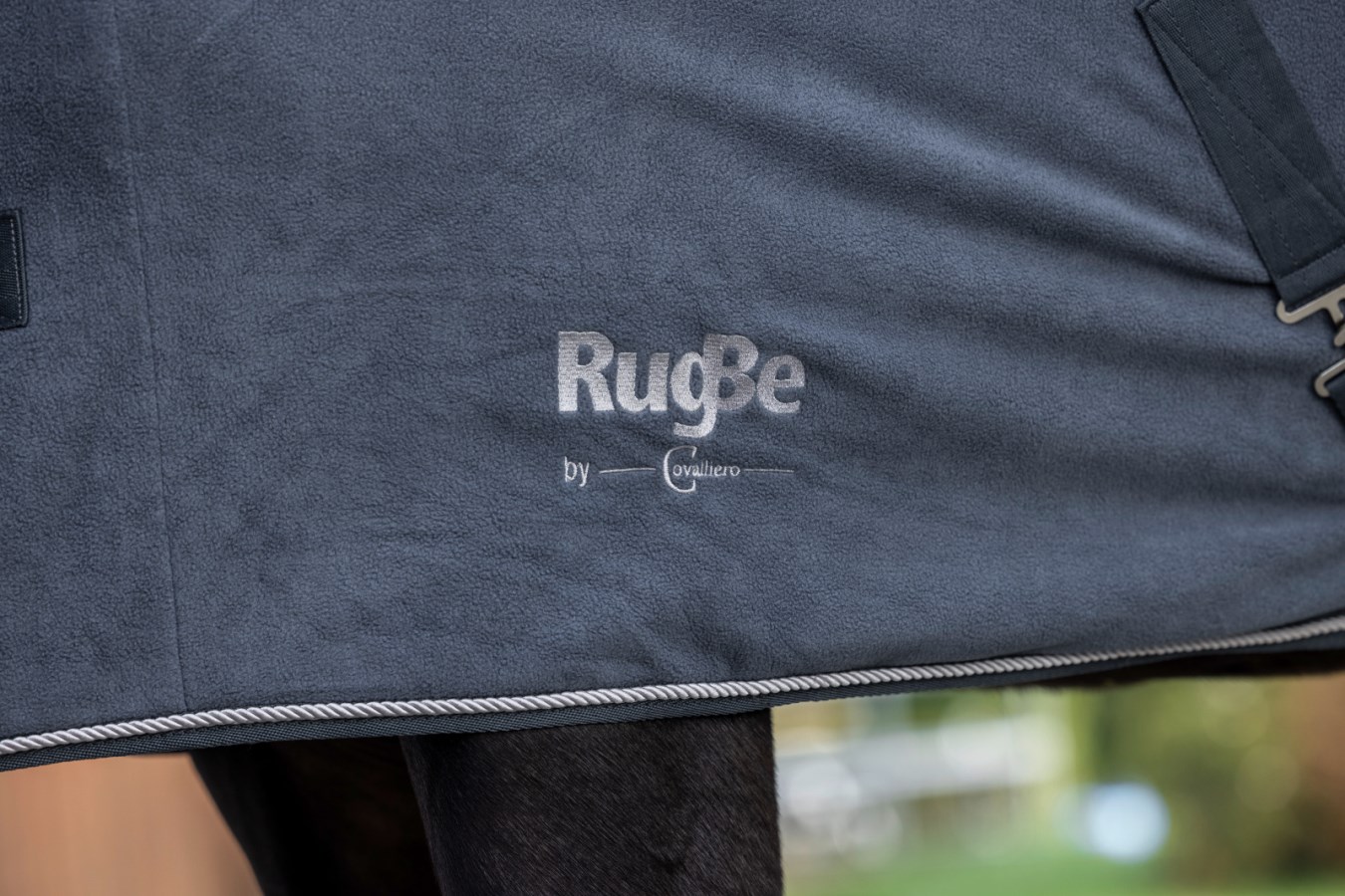 Graphitfarbene Fleecedecke RugBe Royal mit silberner Logo-Stickerei und Zierkordel in der Detailansicht am Pferd.