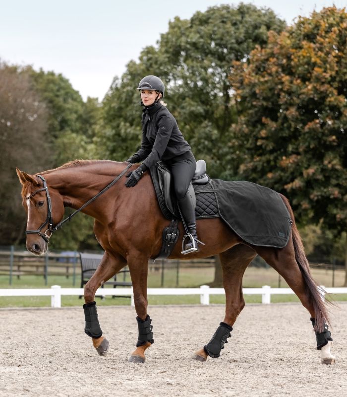 WALDHAUSEN Regen-Nierendecke Salzburg – schwarz Eine Reiterin reitet ein braunes Pferd mit der schwarzen Regen-Nierendecke Salzburg auf einem Sandplatz.