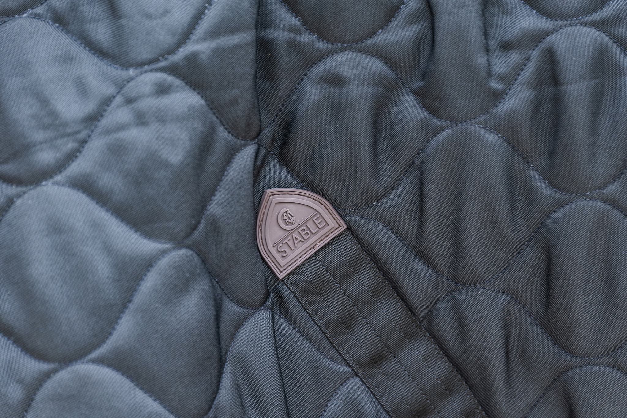 KENTUCKY® HORSEWEAR Stalldecke Comfort 100 g, schwarz Nahaufnahme der schwarzen Stalldecke Comfort mit wellenförmiger Steppung und braunem Markenlogo-Patch.