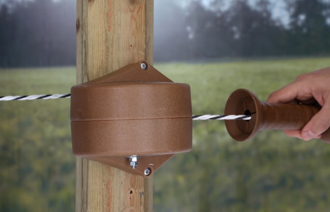 Das braune AKO FLEXIGATE Tor-Set mit Seilaufroller ist an einem Holzpfosten befestigt. Hand zieht am Torgriff.