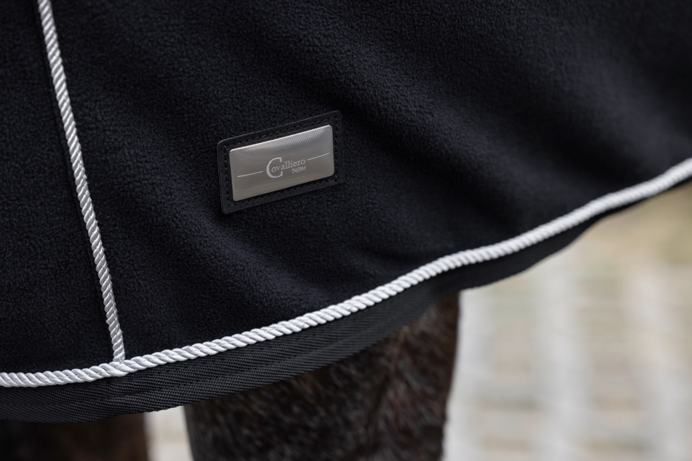 Detail der schwarzen KERBL Fleecedecke CovallieroTherm mit Halsteil, silberner Zierkordel und metallenem Logo-Patch.