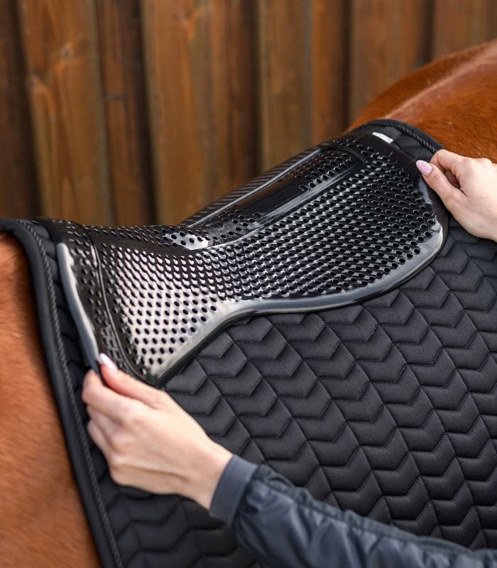 Eine Person legt das schwarze Waldhausen Gel-Pad Basic mit Lochstruktur auf eine gesteppte Satteldecke.