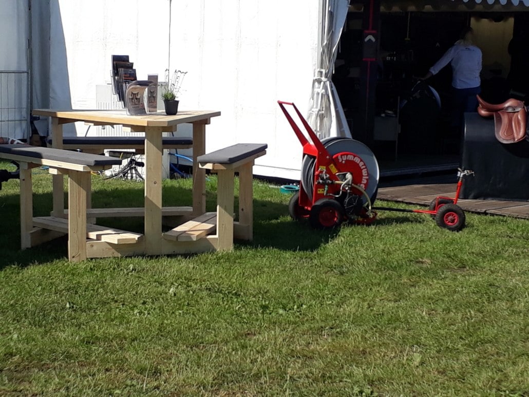 Hölzerne Picknick-Sitzgruppe mit integrierten Bänken und Polstern auf einer Wiese vor einem Messestand