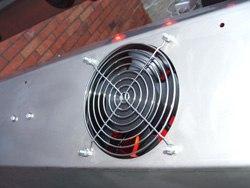 Nahaufnahme eines runden Lüfters mit silbernem Schutzgitter, montiert mit Flügelmuttern auf einem Metallgehäuse