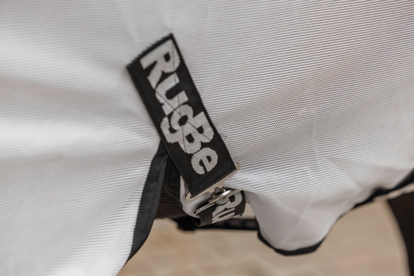 Detailaufnahme der weißen Fliegendecke RugBe SuperFly mit feinmaschigem Gewebe und schwarzem Logo am Verschluss.