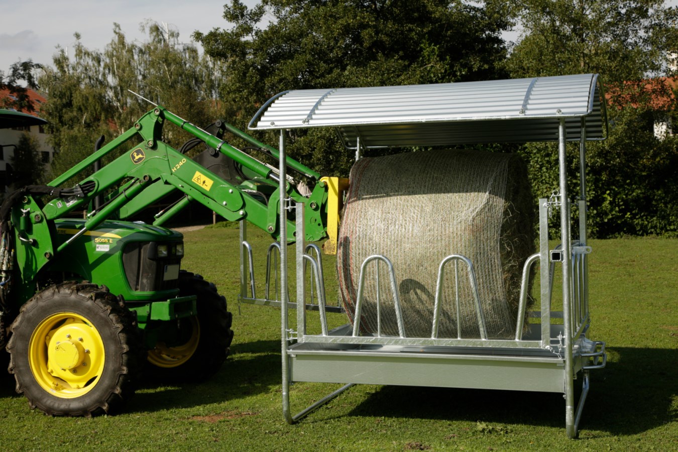 Eine verzinkte Kerbl Viereckraufe mit Palisadenfressgitter und Dach wird von einem Traktor mit einem Heuballen befüllt.
