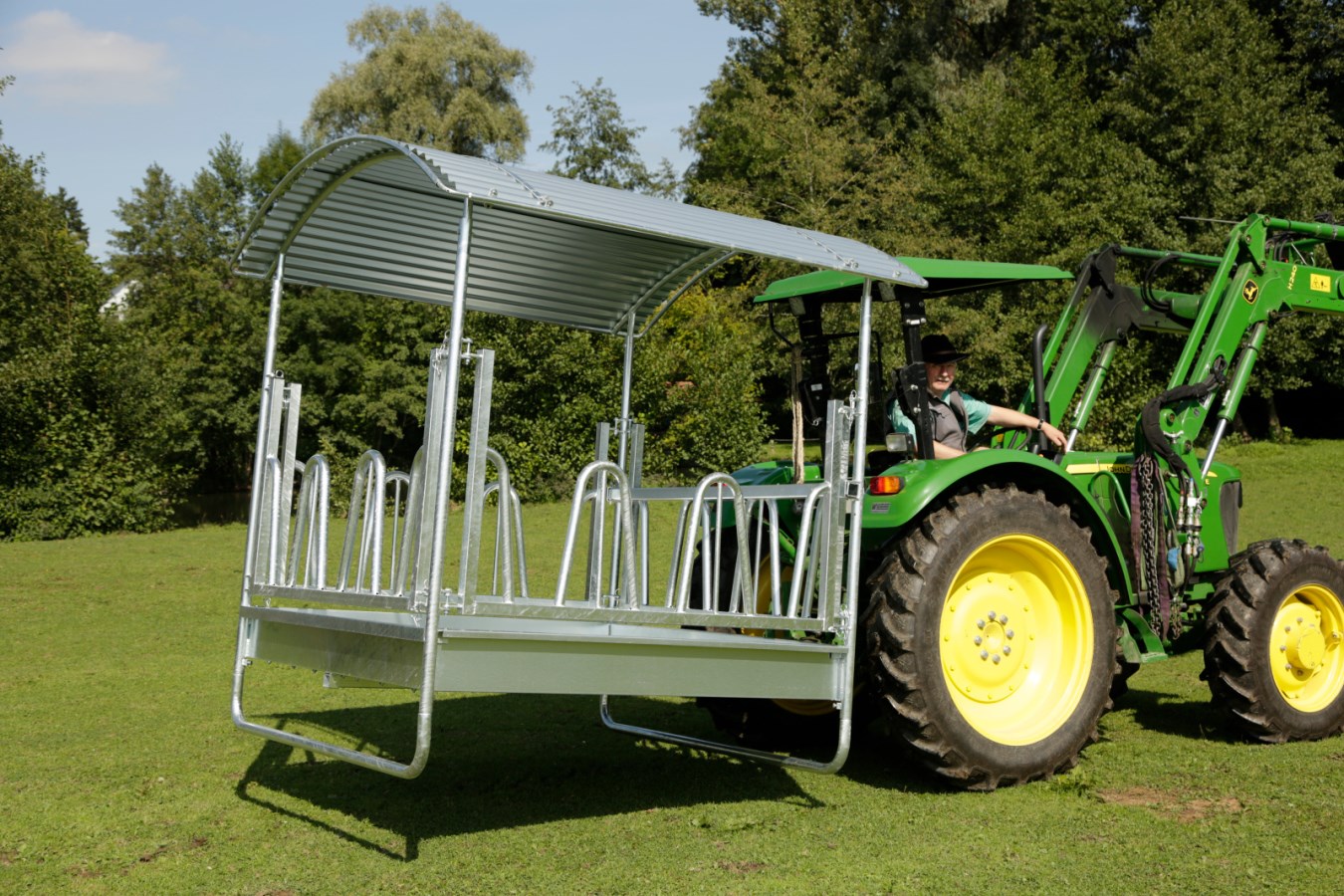 Verzinkte Viereckraufe Standard mit Palisadenfressgitter und Dach wird von einem Traktor auf einer Wiese transportiert.